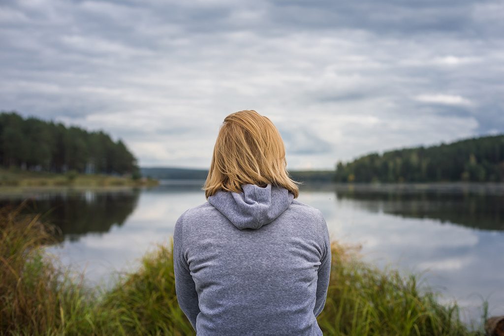 Donna che osserva il lago in silenzio, immagine che rappresenta la riflessione sull'identità, i cambiamenti della vita e il benessere psicologico.